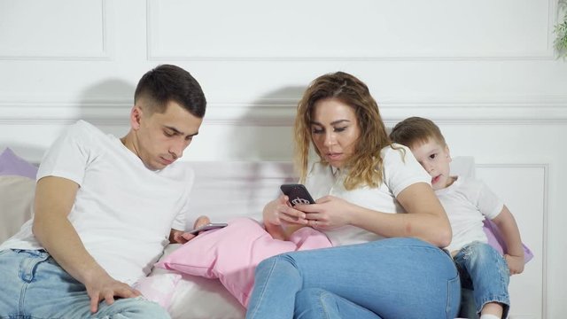 Mom and dad are looking in their mobile phones not paying attention to their child laying in bed. Sad little boy wants to play and pulls mom's hand. Escape of reality, gadgets addiction.