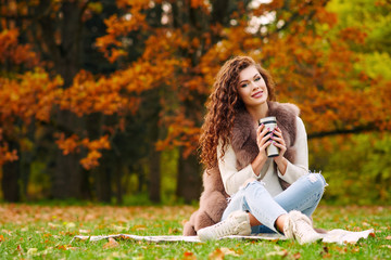 Slender beautiful young woman in a fur vest is resting in the autumn in the park and sitting on the lawn on the lawn and drinking tea from a thermocup