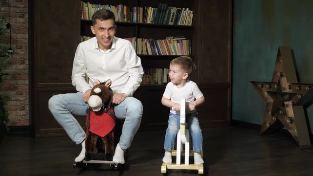 Happy family young dad and little cute son are playing together riding on toy horses rocking chair in living room at home. Family time.
