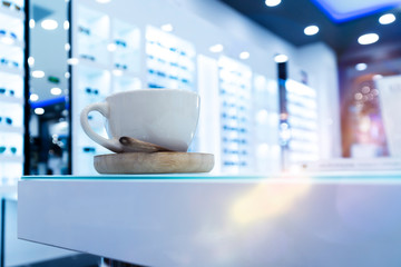 Closeup photo of cup with saucer standing on the glass and white  table in the sunglasses shop with blurred background.