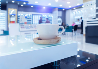 Closeup photo of cup with saucer standing on the glass and white  table in the sunglasses shop with blurred background.