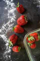 Ripe, red strawberries with powdered icing sugar and glass container on dark background. Top view.