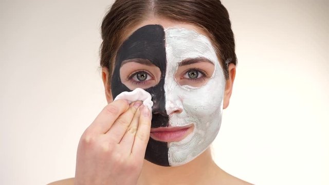 Woman with clay carbo black mask on one half face and white mud on second half. Girl removing cosmetic scrab with cotton swab. Female taking care of oily complexion. Skincare