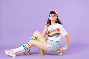 Naklejka premium Tender teen girl in vivid clothes, eyeglasses sitting on yellow skateboard blowing sending air kiss isolated on violet pastel background. People sincere emotions lifestyle concept. Mock up copy space.