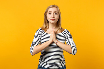 Portrait of pleading young woman in striped clothes folded her hands and praying isolated on yellow orange wall background in studio. People sincere emotions, lifestyle concept. Mock up copy space.