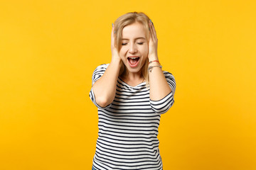 Screaming young woman in striped clothes keeping eyes closed, putting hands on head isolated on yellow orange wall background in studio. People sincere emotions, lifestyle concept. Mock up copy space.