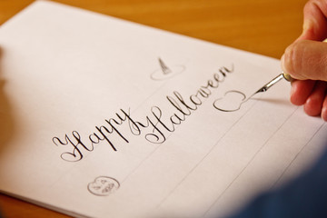 hand writes with the inky pen the happy halloween on a white paper sheet with stripes. stationery on desk close up top view. spelling lessons and caligraphy exercises. Template, layout, background