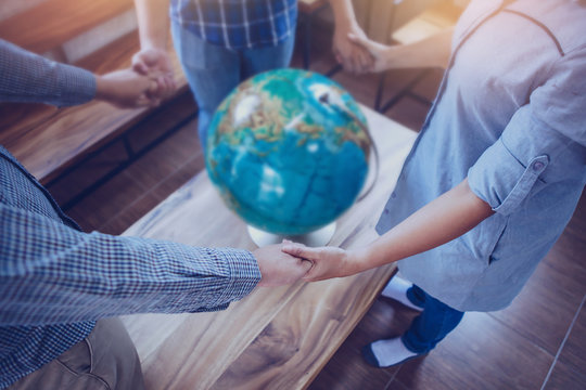 Three People Standing And Holding Hands Each Other While Pray To God For The World With Blurred World Globe On Wooden Table, Christian Background For Great Commission Or Earth Day Concept.