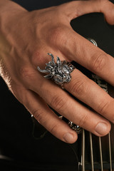 Cropped closeup shot of man's hand, touching headstock of guitar. The guy is wearing massive signet-ring in view of demon Baphomet. The man is wearing black clothes, posing against dark background.