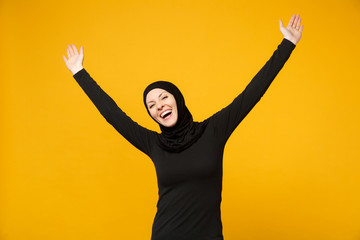 Overjoyed young arabian muslim woman in hijab black clothes doing winner gesture, say Yes isolated on yellow wall background, studio portrait. People religious lifestyle concept. Mock up copy space.