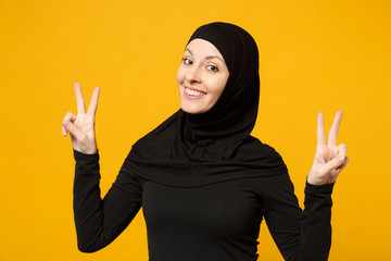 Smiling beautiful young arabian muslim woman in hijab black clothes showing victory sign isolated on yellow background, studio portrait. People religious Islam lifestyle concept. Mock up copy space.