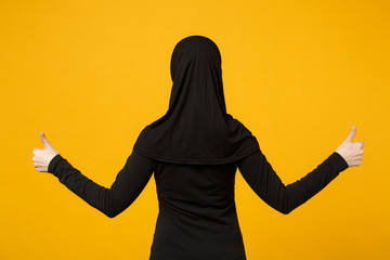 Back rear view image of young arabian muslim woman in hijab black clothes showing thumbs up isolated on yellow wall background, studio portrait. People religious lifestyle concept. Mock up copy space.