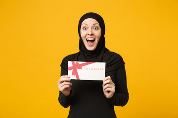 Overjoyed young arabian muslim woman in hijab black clothes hold in hands gift certificate isolated on yellow wall background, studio portrait. People religious lifestyle concept. Mock up copy space.