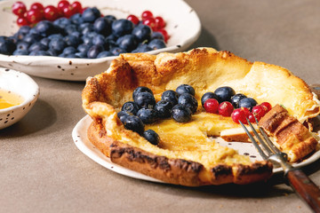 Started eaten fresh baked Dutch baby pancake in ceramic plate with blackberry and red currant berries, bowl of honey, jug of cream, vintage cutlery over beige kitchen table. Close up