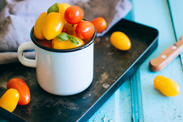 Red and yellow small cherry tomatoes, and green basil in an enamel mug on a black tray
