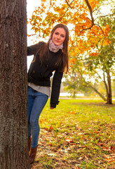 Beautiful girl in autumn park, hiding behind a tree and smiling at the camera. Gorgeous young woman outdoors, in a warm sunny day. Full length body shot in natural light, retouched, vibrant colors