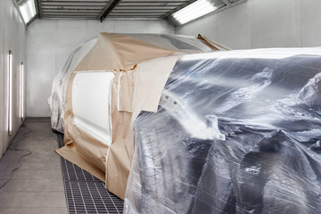 A large white car is completely covered in paper and adhesive tape to protect against splash during painting and repair after an accident in a workshop for body repair of vehicles with bright lighting