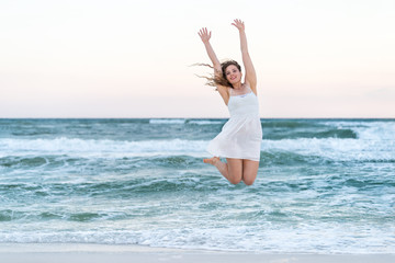 Young happy smiling woman in white dress on beach sunset in Florida panhandle with ocean waves jumping mid-air