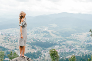 Naklejka premium Young beautiful barefoot blonde girl with long hair in summer dress standing on top of conquered mountain at stone and enjoying fabulous landscape scenic view with mountains and village in valley