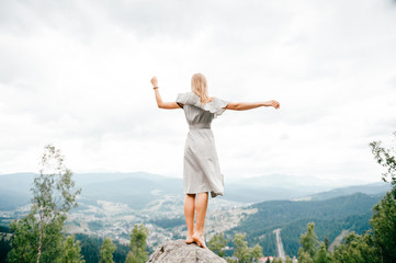 Fototapeta na wymiar Young beautiful barefoot blonde girl with long hair in summer dress standing on top of conquered mountain at stone and enjoying fabulous landscape scenic view with mountains and village in valley
