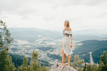 Naklejka premium Young beautiful barefoot blonde girl with long hair in summer dress standing on top of conquered mountain at stone and enjoying fabulous landscape scenic view with mountains and village in valley