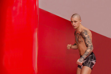 Tattooed young MMA fighter on ready to strike a punching bag standing in front of it. Mixed martial arts training in a gym. Preparations before fight. Bold man with tattoos fights. Sport, lifestyle.
