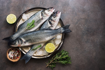 Flat lay fresh fish sea bass in a plate with ice cubes, rosemary and lime on a dark rustic...