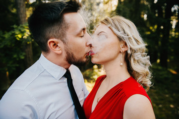 Closeup of couple of lovers kissing with smoke outdoor.  Businessman and young blonde girl with red lips, curly hair and red dress with decolette. Smokers in forest in summer sunny day. Sensual moment