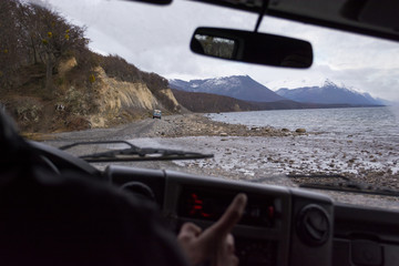 Inside view from the interior of a vehicle to the front window, with a dirt rocky route on the background next to a lake. The driver is in a blur pointing out and the interior is dark, with copy space