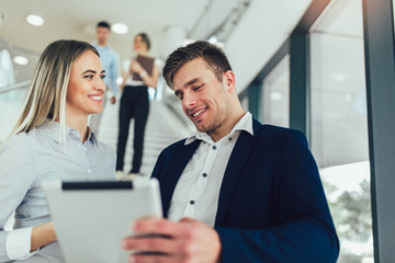 Beautiful business woman and man their holding a tablet in hands and smiling at the camera. In the background are business people.