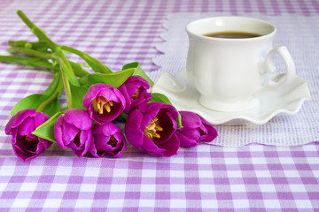 White porcelain cup with tea or coffee on saucer with wavy edge and a bouquet of purple tulips on a checkered tablecloth. Greeting card with copy space.