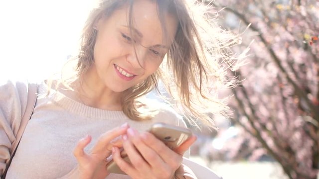 Close portrait of a young woman with a smartphone in her hands. The girl smiles leafing through the photo in her phone. Portrait on the background of flowering spring trees