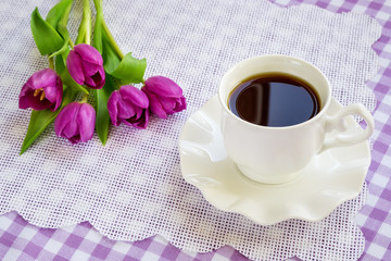 White porcelain cup of tea or coffee on saucer with wavy edge and a bouquet of purple tulips on a checkered tablecloth. Greeting card with copy space.