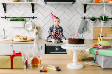 childhood, people and celebration concept - happy baby girl in pink birthday hat sitting in the kitchen on birthday party at home. Table with birthday cake, presents, air balloons and party horns