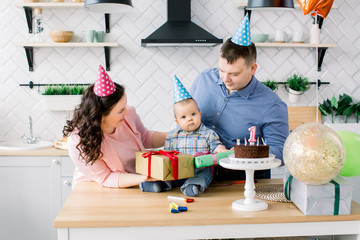 Mother and father in birthday hats celebrating first birthday of baby boy at home kitchen. Happy family celebrating birthday with a cake, present boxes, party horns and balloons