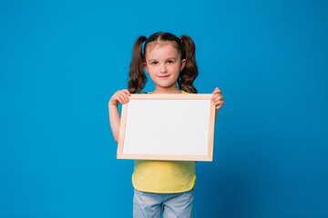 baby brunette girl in yellow t-shirt on blue background holding white sheet. space for text. children's emotions