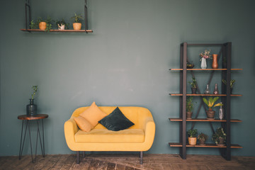 Interior design room or studio. Furniture against a green wall and wooden floor - a wooden table with a vase, a yellow sofa with cushions, a wardrobe with flowers and plants, a hanging shelf