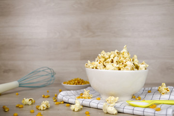 Popcorn in white bowl with pastel spoon, salt, hand mixer and corn seeds on wood background....