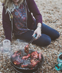 A woman sitting on a stony beach flipping chicken on a barbecue with tongs