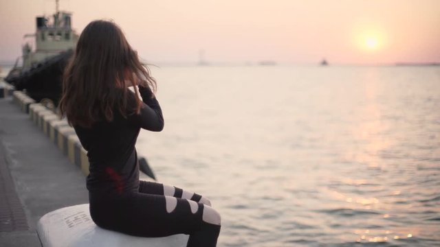 Young stylish woman with riding roller skates near sea port during sunset and using her smartphone, makes selfie.