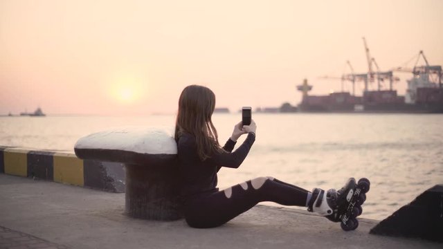 Young stylish woman with riding roller skates near sea port during sunset and using her smartphone, makes selfie.