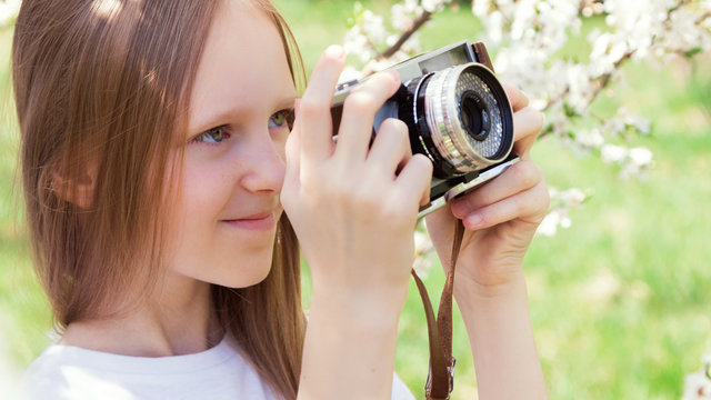 A beautiful little thin girl teenager walks through a flowered garden and photographs the plants on a retro-photographic camera. Spring. The game is in the open air. Summer educational camp.