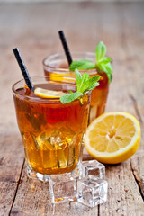 Traditional iced tea with lemon, mint leaves and ice cubes in two glasses on rustic wooden table.