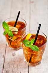 Traditional iced tea with lemon, mint leaves and ice cubes in two glasses on rustic wooden table.