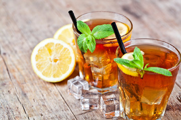 Traditional iced tea with lemon, mint leaves and ice cubes in two glasses on rustic wooden table background.