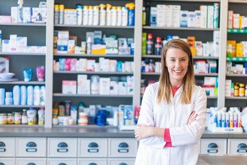 Portrait of a smiling pharmacist with arms crossed at modern pharmacy. Beautiful woman wearing in...