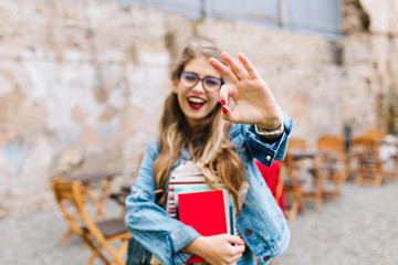 Charming female student with books showing her excitement and holding up her thumb. Outdoor portrait of young lovely lady with smiling facial expression.