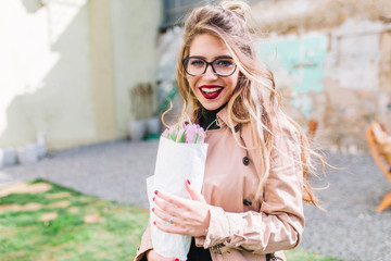 Outdoor close-up portrait of birthday girl holding beautiful purple tulips in paper wrapper and looking with grateful smile. Young fair-haired lady on the date in cafe carrying flowers from boyfriend