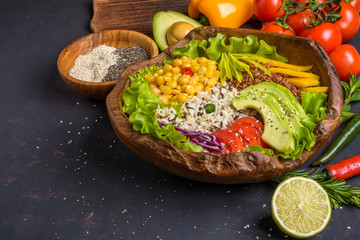Buddha bowl with chickpea, avocado, wild rice, quinoa seeds, bell pepper, tomatoes, greens, cabbage, lettuce on shabby dark stone table and wooden bowl with chia and sesame seeds.