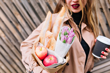Lovely girl with red lips holding a grocery bag, flowers and cappuccino isolated on blur wooden background. Stylish woman posing after shopping in bakery, focus is on paper bag in front. Tasty meal